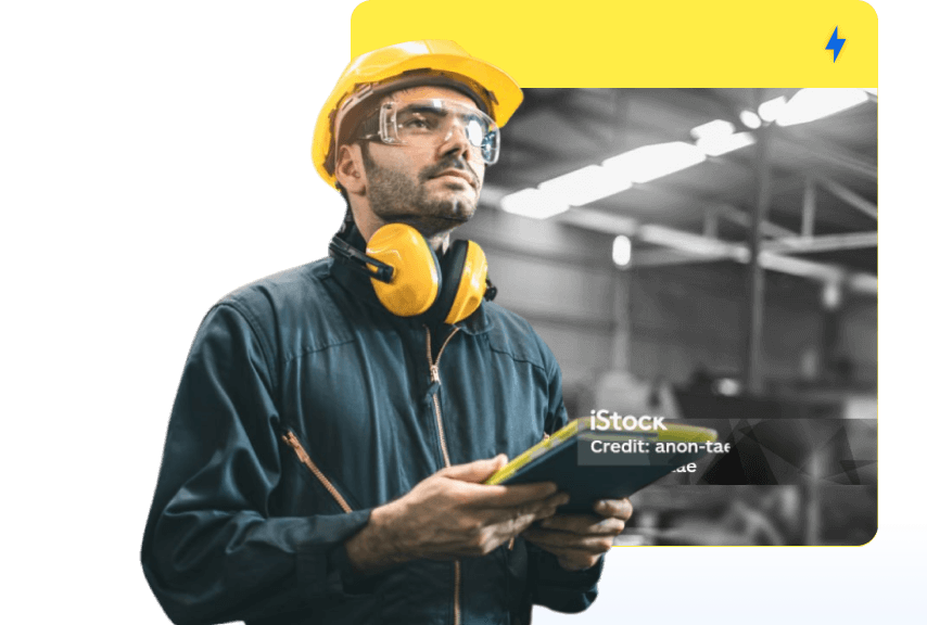 A man in a yellow hard hat holding a tablet in front of an image of a manufacturing facility.